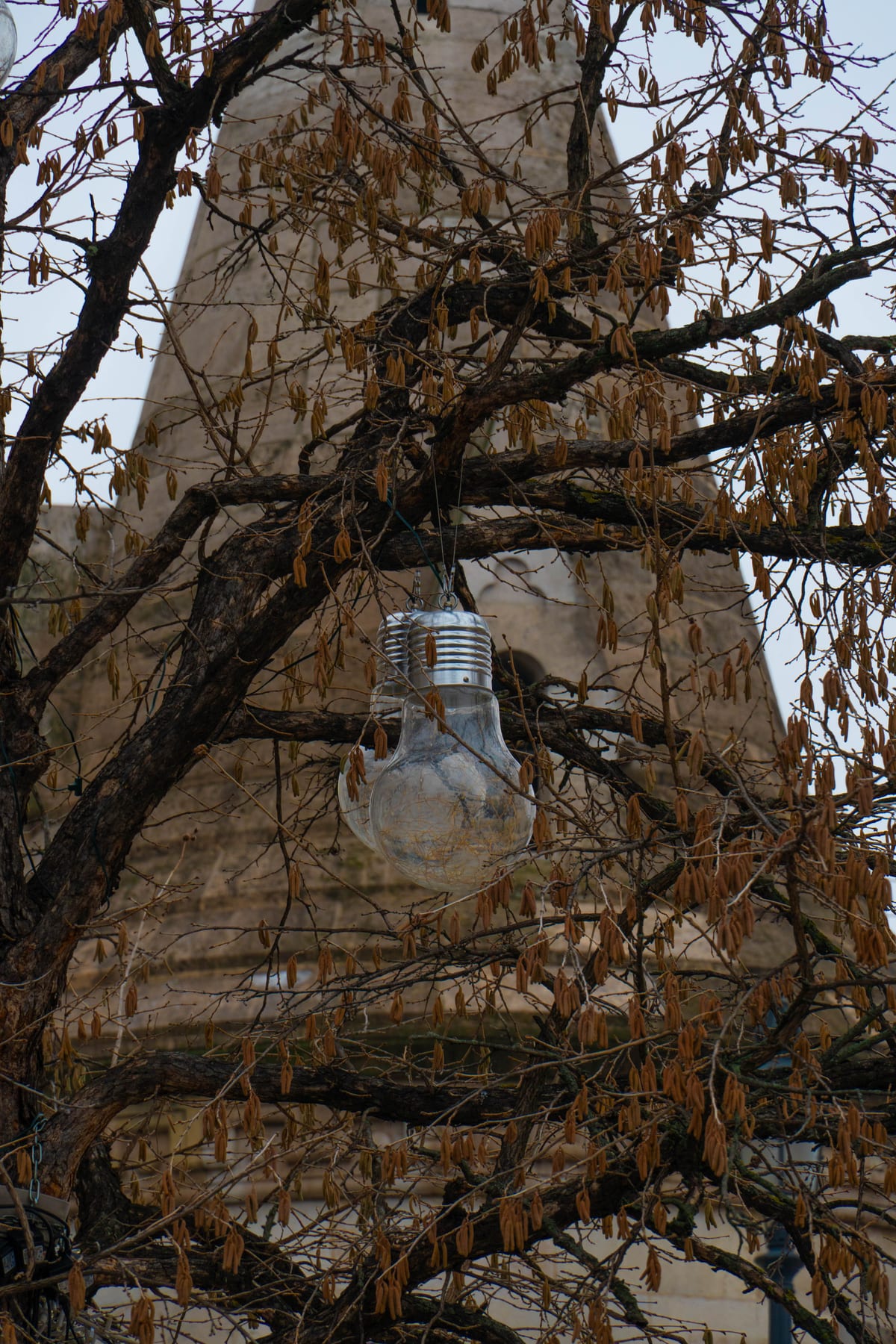 Giant light bulb hanging in a bare tree in front of a stone tower